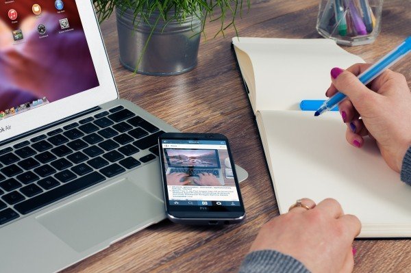 woman-sitting-at-table-writing-in-notepad-with-laptop-and-mobile-phone-on-wooden-table